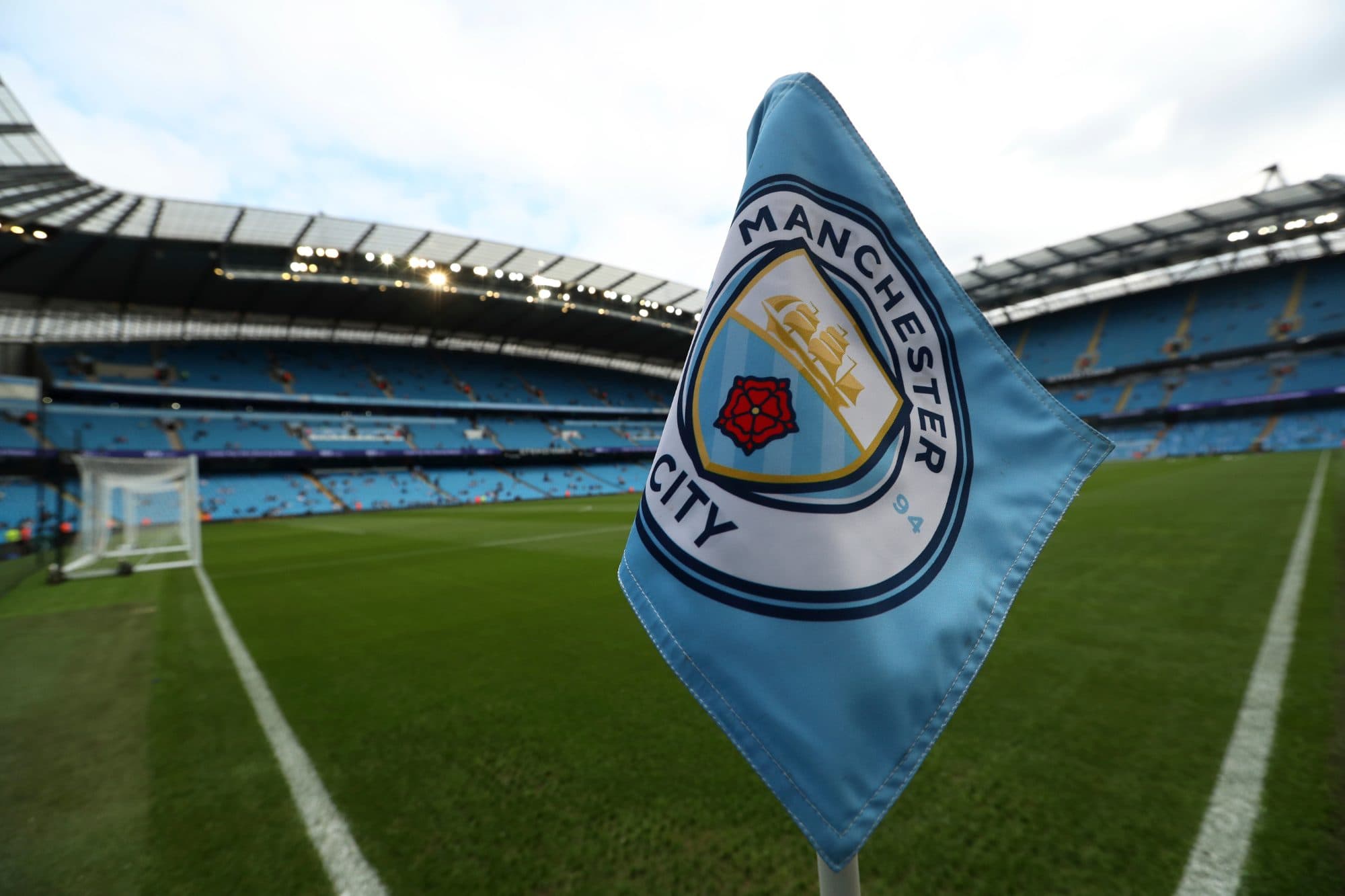 A close-up shot of a sky-blue corner flag featuring the official Manchester City FC crest set at the edge of a football pitch. In the blurred background, the stadium stands with blue seating and a curved roof structure are visible under an overcast s